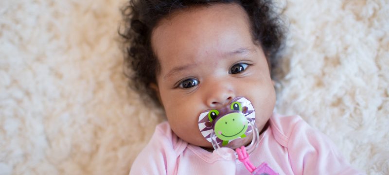 Baby laying on rug looking up at camera with pacifier in her mouth